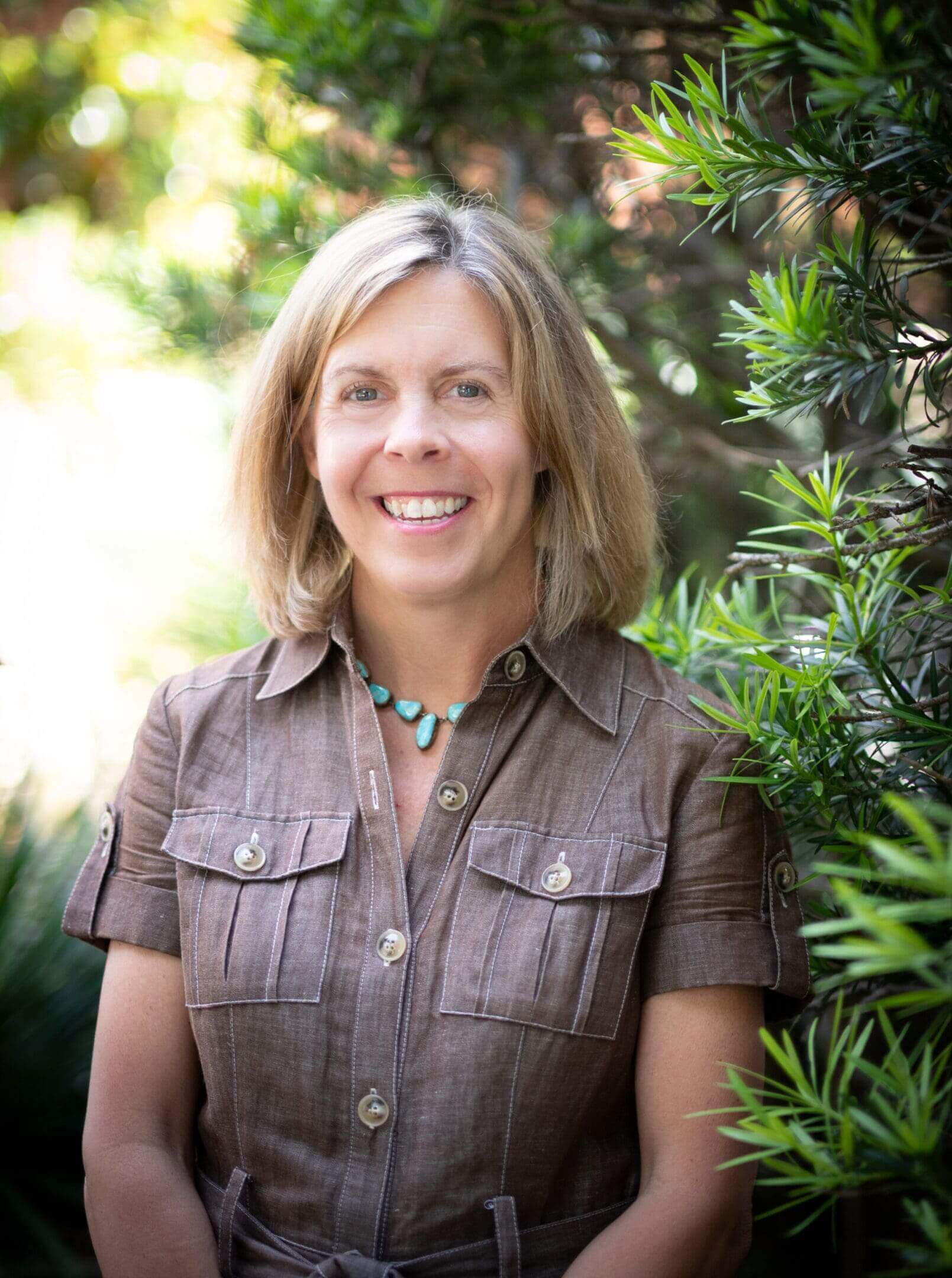 Smiling woman outdoors with greenery in the background.