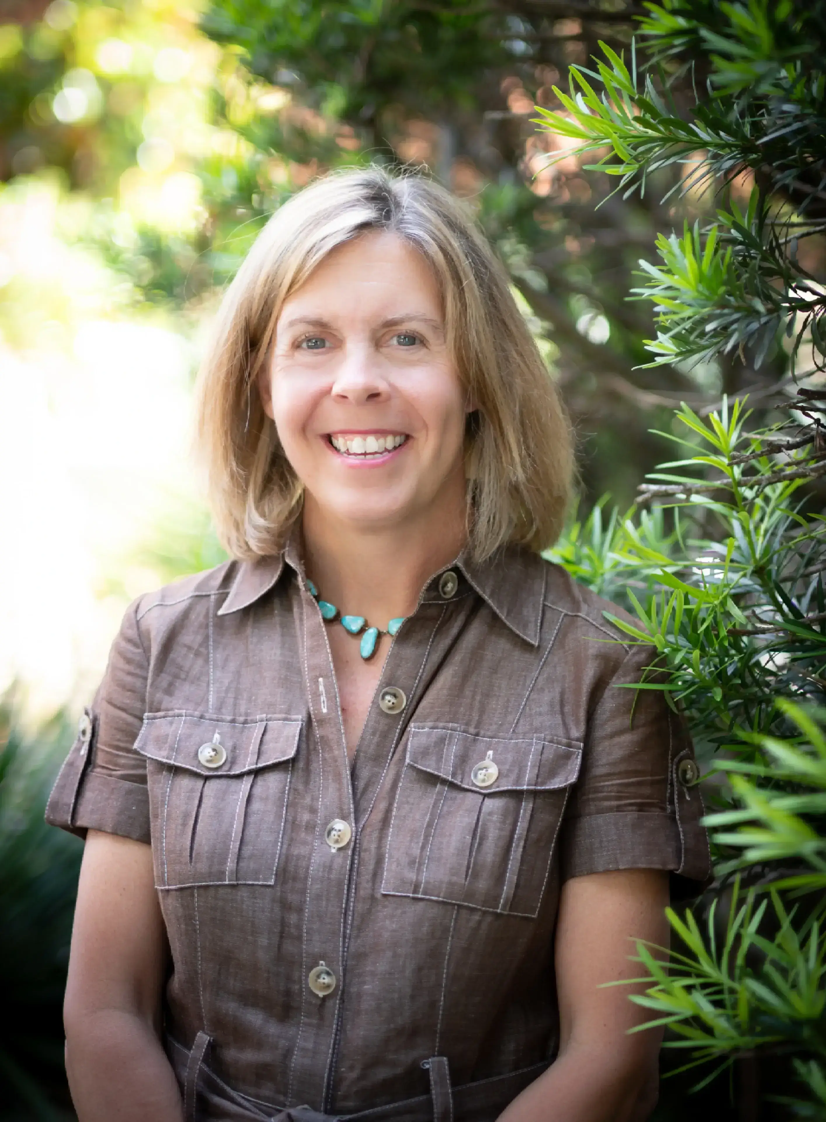 Smiling woman seated near green foliage.